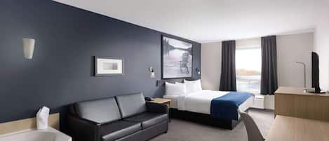 Habitación de hotel con una pared azul oscuro, un sofá de cuero negro, cama con manta azul, escritorio de madera, bañera blanca y un ventanal con cortinas.