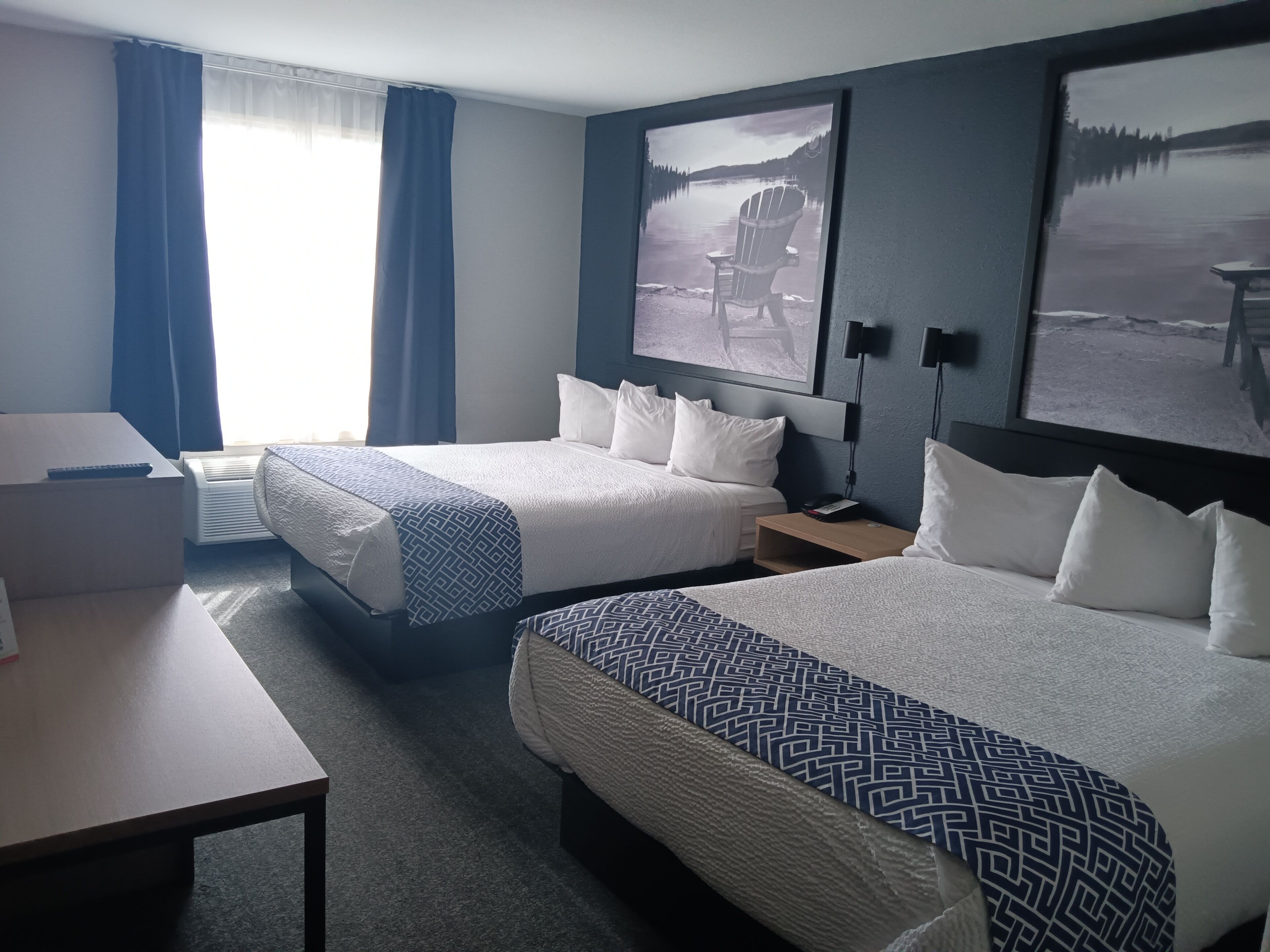 Habitación de hotel con dos camas, un escritorio y una silla. Hay dos cuadros en la pared, una ventana con cortinas y un equipo de aire acondicionado.