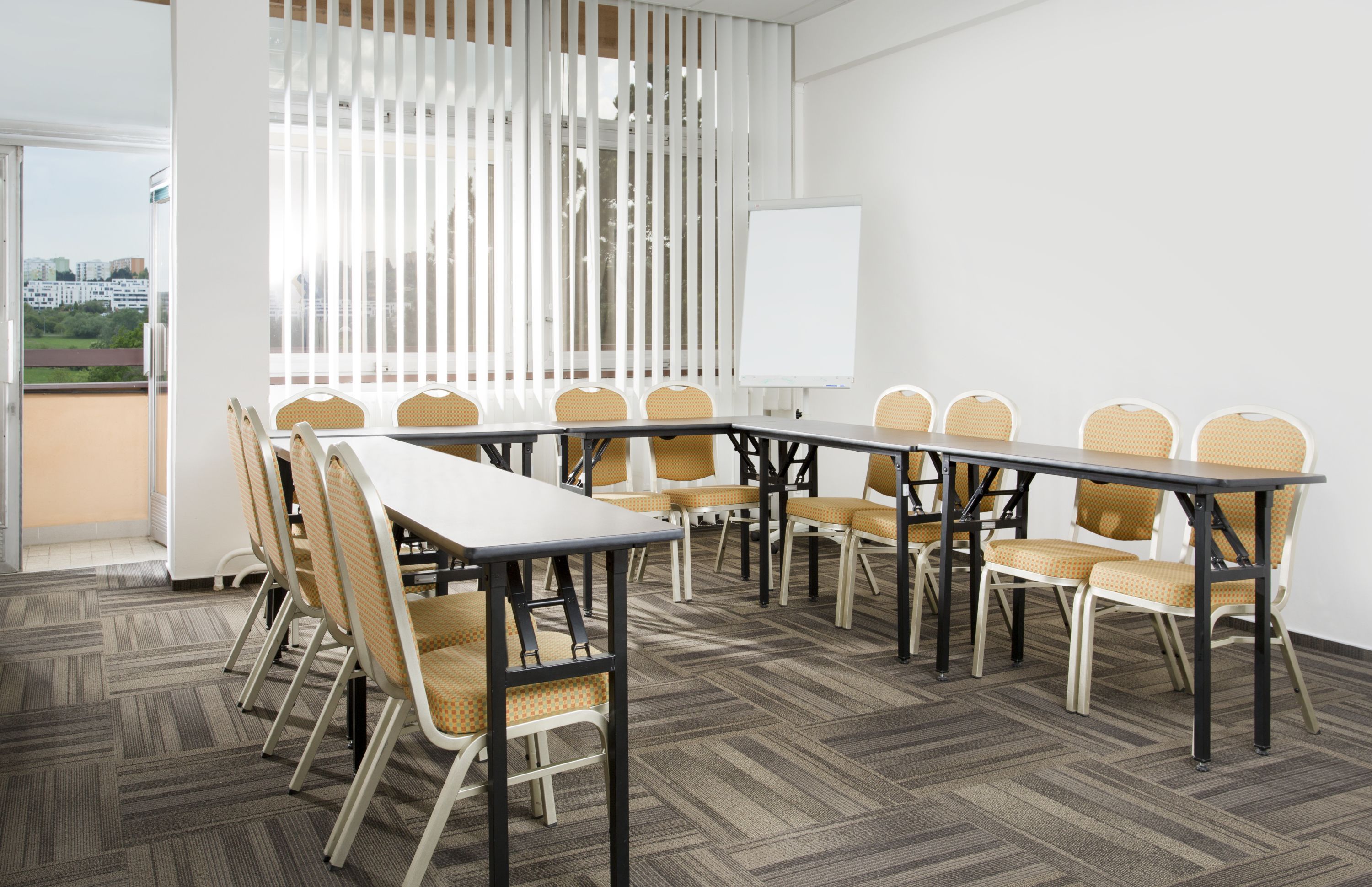 A conference room with a long table and chairs, a whiteboard, and a view through a window with blinds.