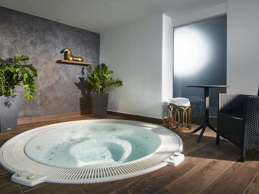 A modern bathroom with a freestanding bathtub, two potted plants, a small round table with a chair, and a decorative horse sculpture on the wall.