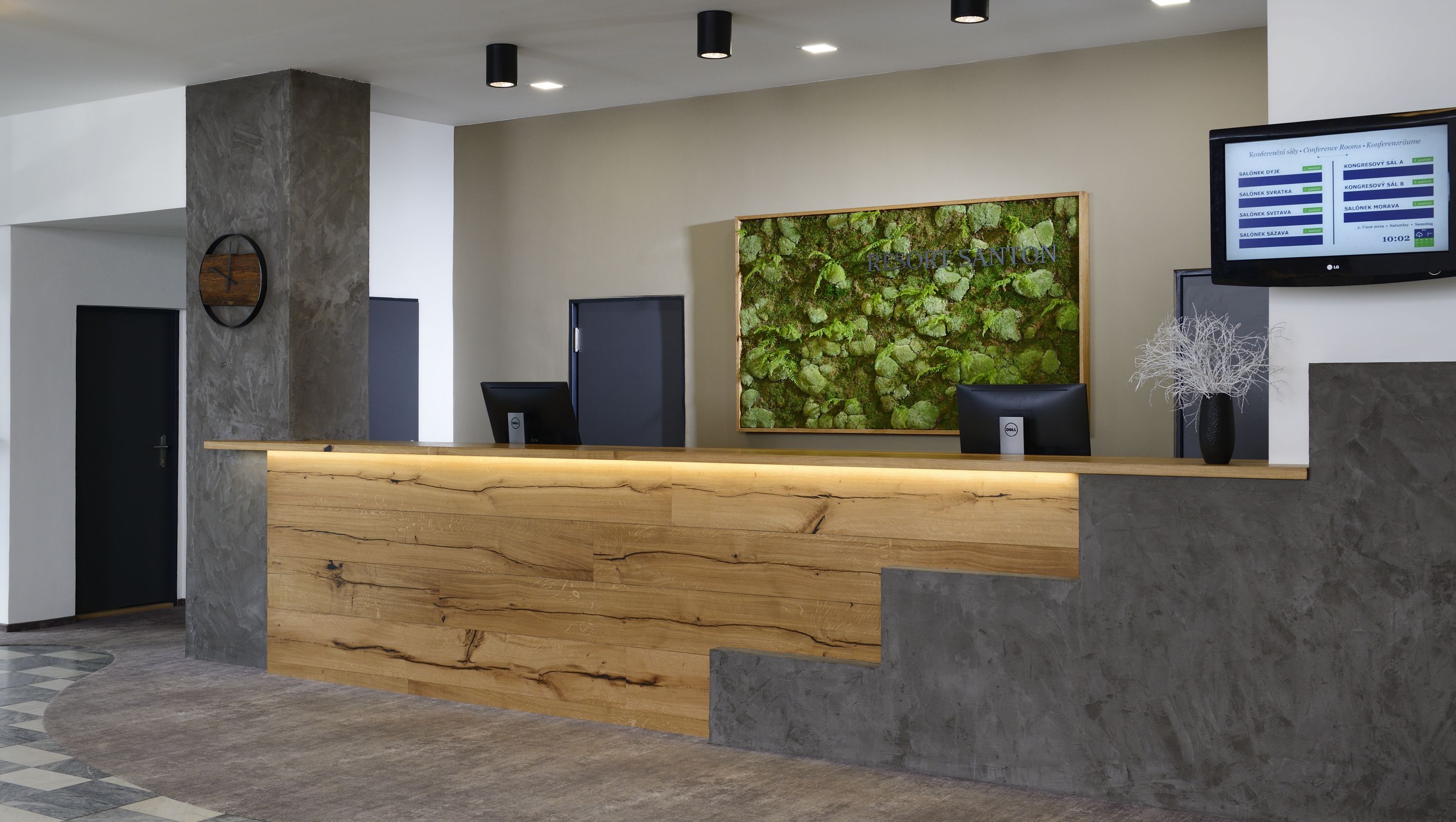 A reception desk with a wooden counter, a large moss wall, and a digital screen displaying conference room information.