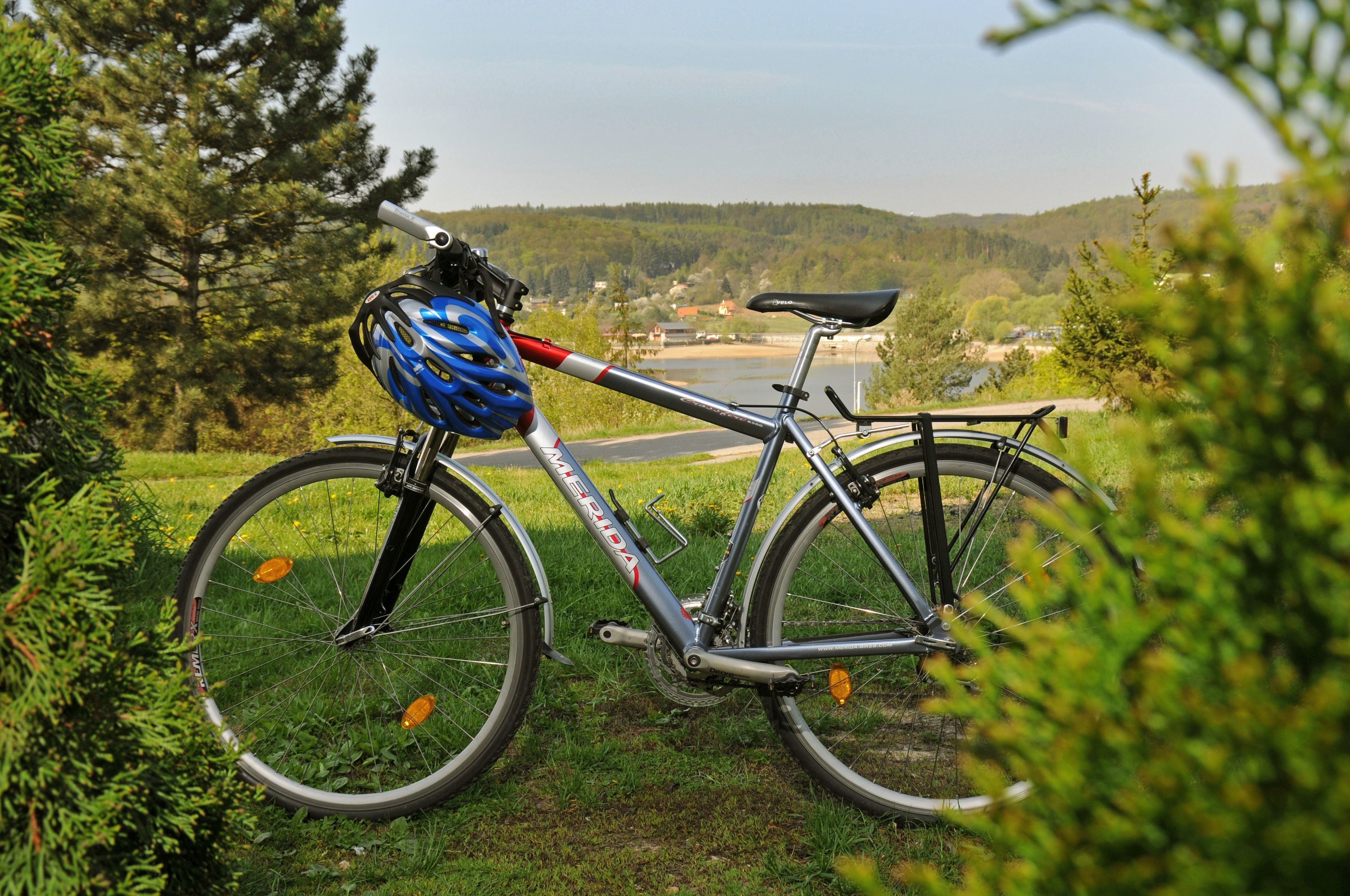 A bicycle with a blue helmet and a water bottle, parked on grass near a lake with trees and hills in the background.