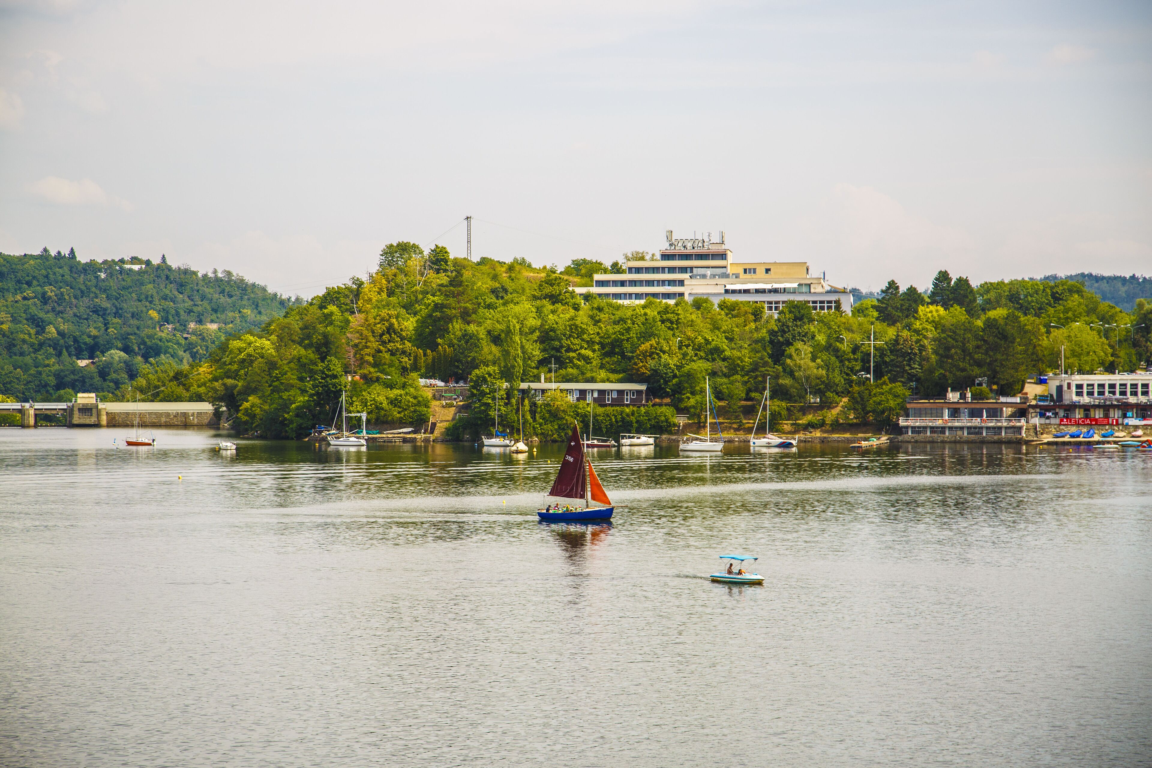 Un lago tranquilo con barcos, un velero en primer plano y un edificio con un letrero en una colina al fondo.