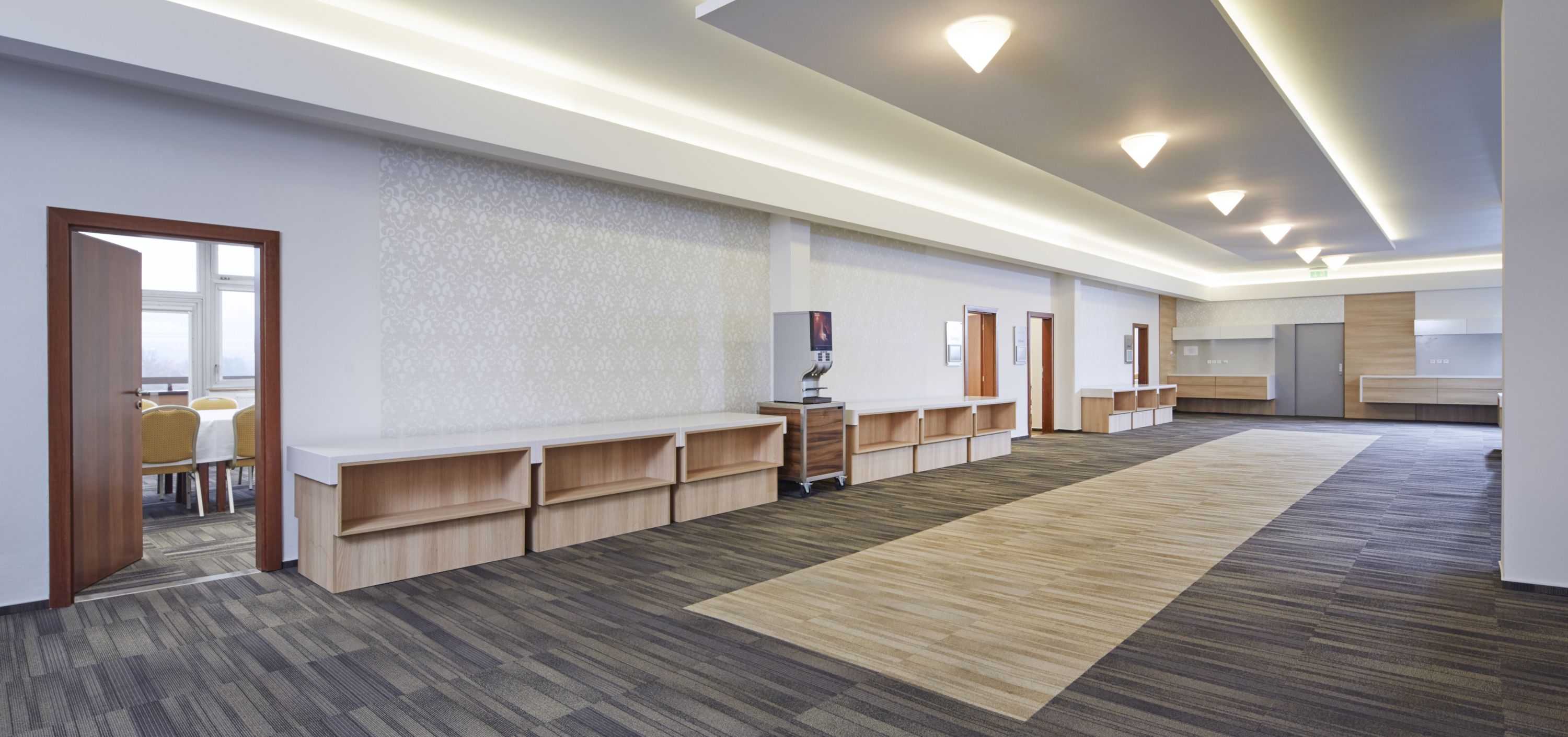 A spacious hotel lobby with a long reception desk, wooden shelving units, and a patterned carpet.
