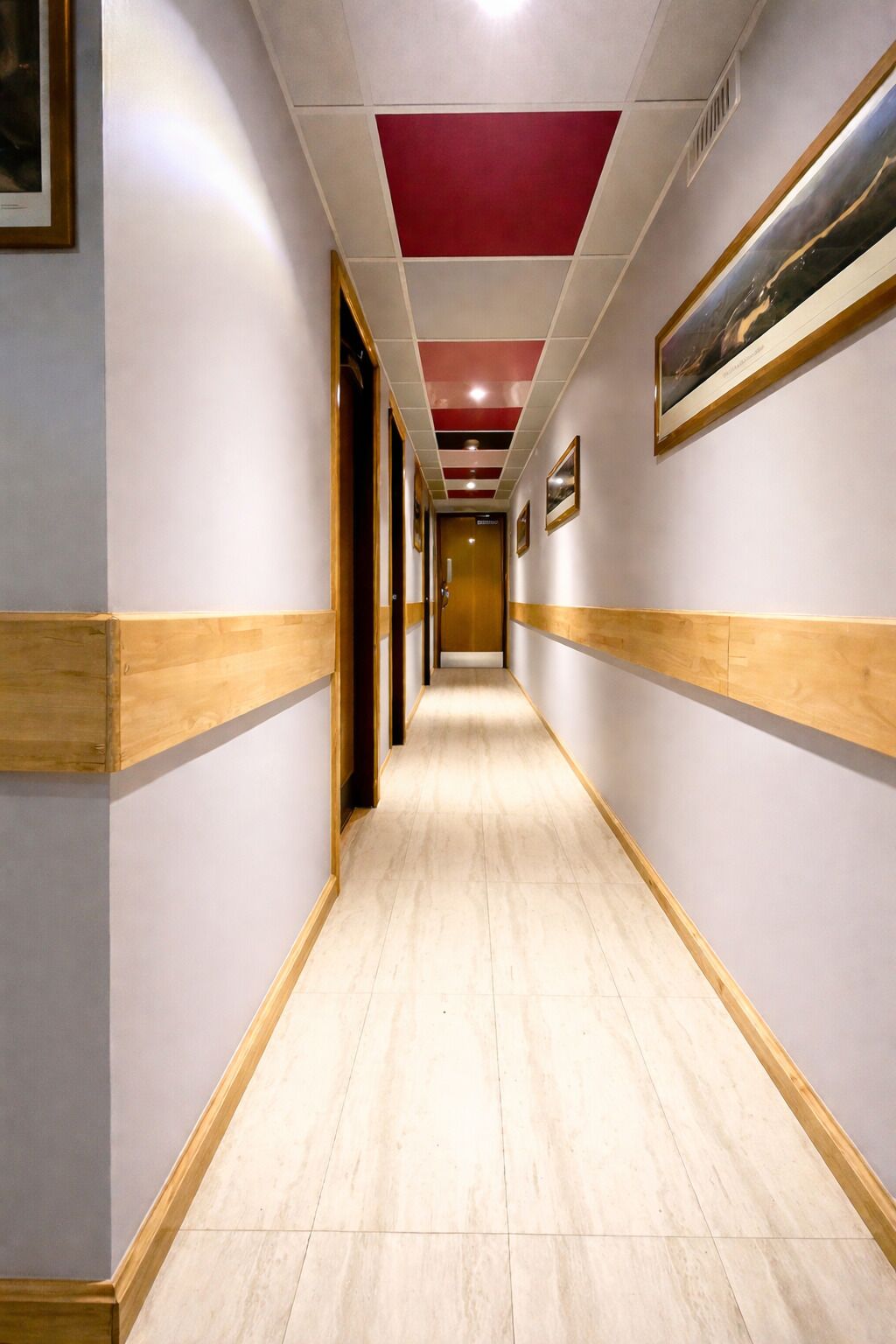 A hotel hallway with a wooden floor, white walls, and a red ceiling panel.