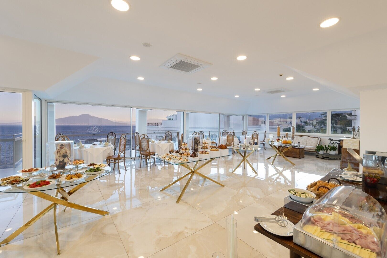 A spacious room with multiple tables set for dining, large windows offering a view of the sea, and a balcony with a view of the city.