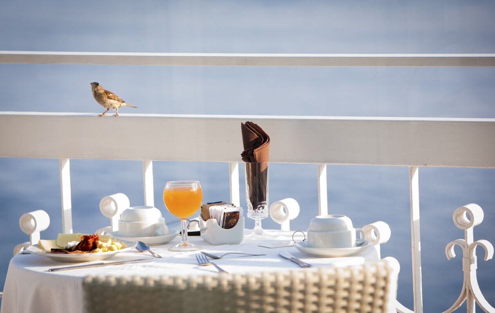 A table set with breakfast items, a glass of orange juice, and a bird on the railing.