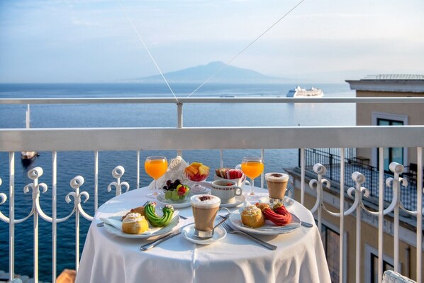 A table set with breakfast items, including fruit, pastries, and beverages, overlooking a body of water with a distant mountain and a cruise ship.