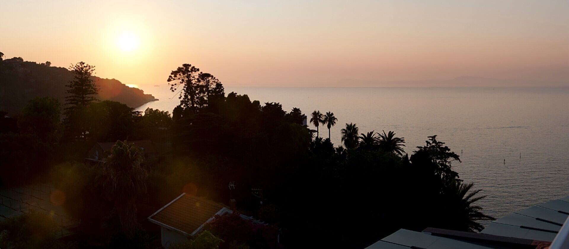 Vista de un atardecer costero con una casa y árboles en primer plano.