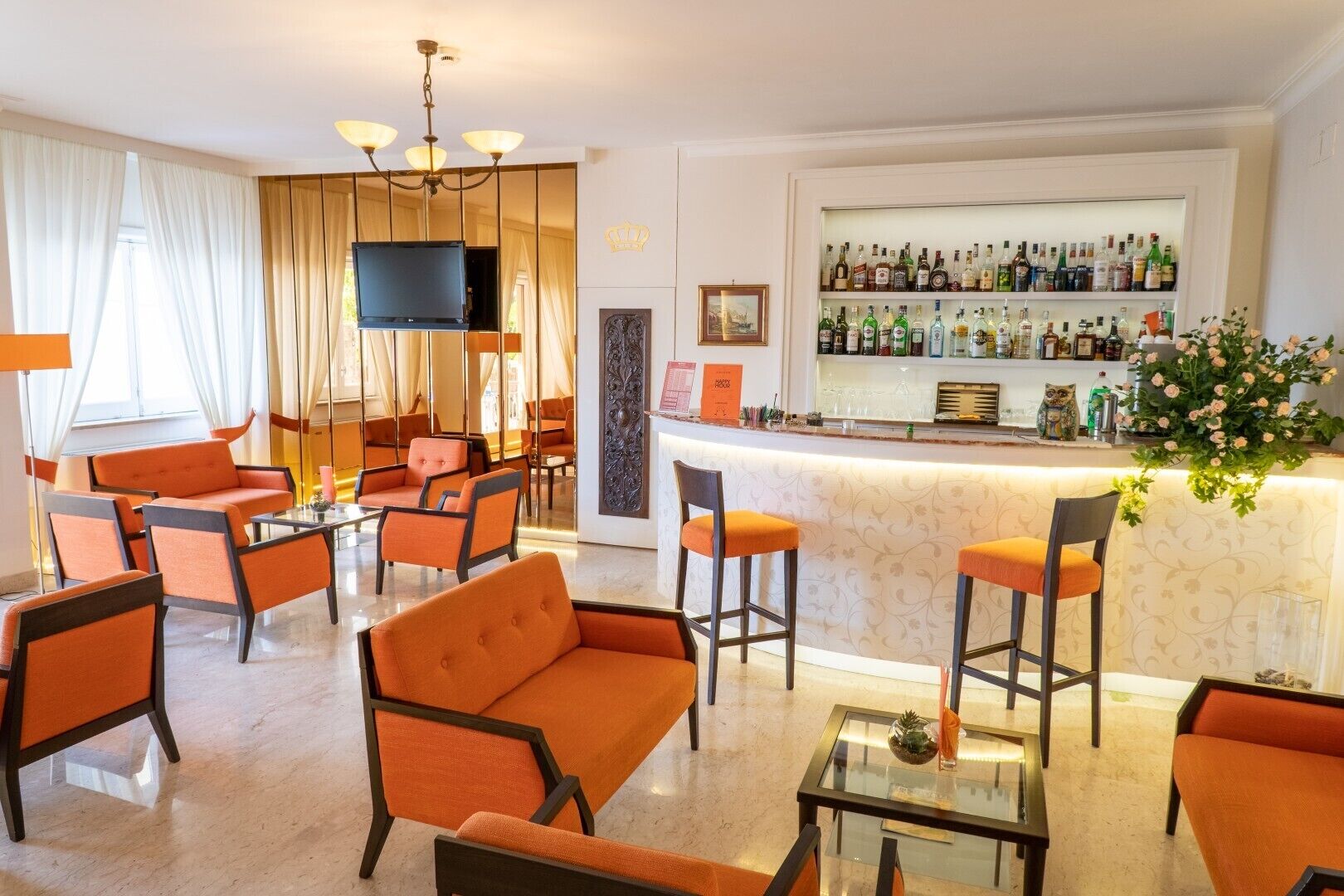 Eine Hotelhalle mit orangefarbenen Sesseln, einer Bar mit verschiedenen Spirituosenflaschen, einem Flachbildfernseher und einer Wanddekoration.