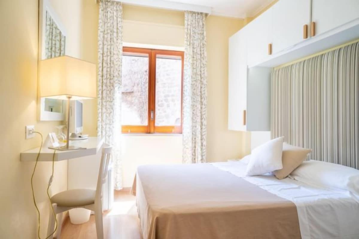 Ein Schlafzimmer mit einem Bett, einem Schreibtisch, einem Stuhl, einem Fenster mit Vorhängen und einem Nachttisch.