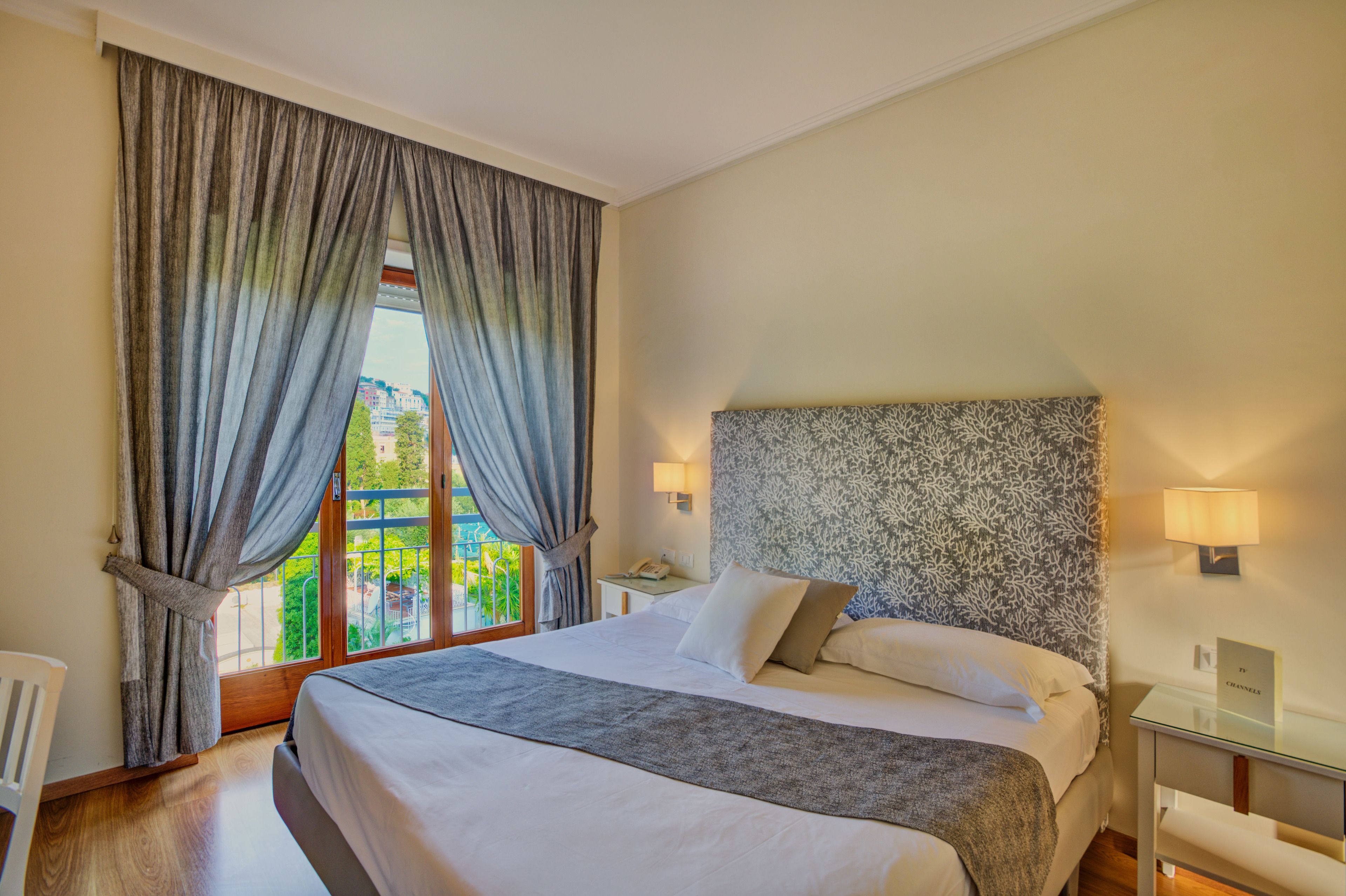 Ein Hotelzimmer mit einem großen Bett, einem gemusterten Kopfteil, zwei Nachttischlampen, Blick auf Grün und einem Fenster mit Vorhängen.