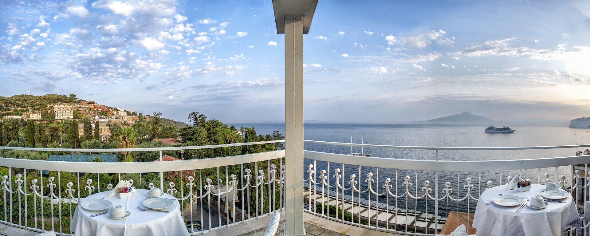 Un balcón con mesa lista para comer, con vista a un paisaje costero que incluye una montaña y un crucero en la distancia.