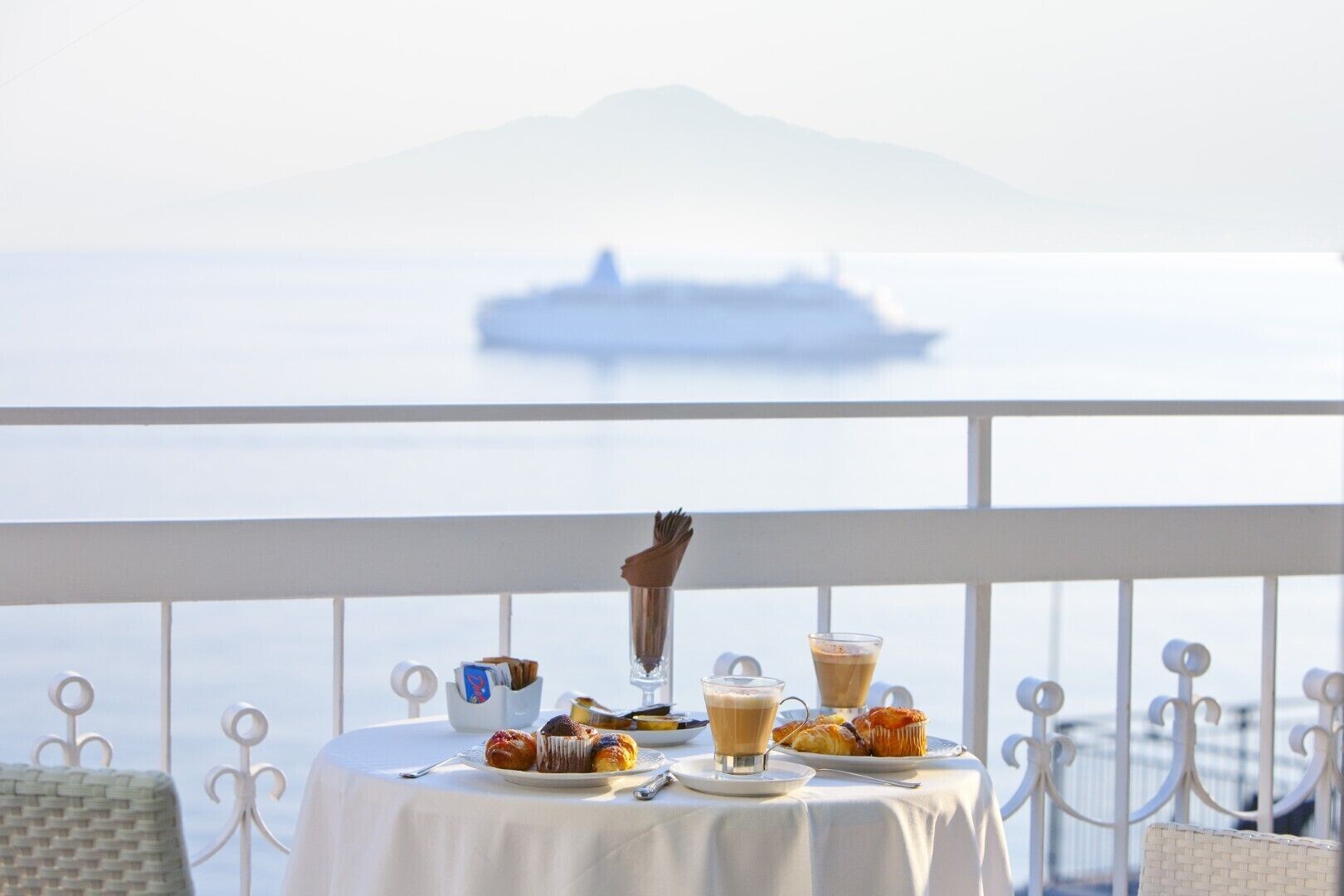 A table with pastries and coffee on a balcony overlooking a calm sea with a ship in the distance.