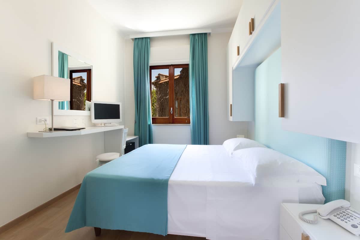 Een hotelkamer met een bed, een bureau met een computer, een telefoon en een raam met gordijnen.