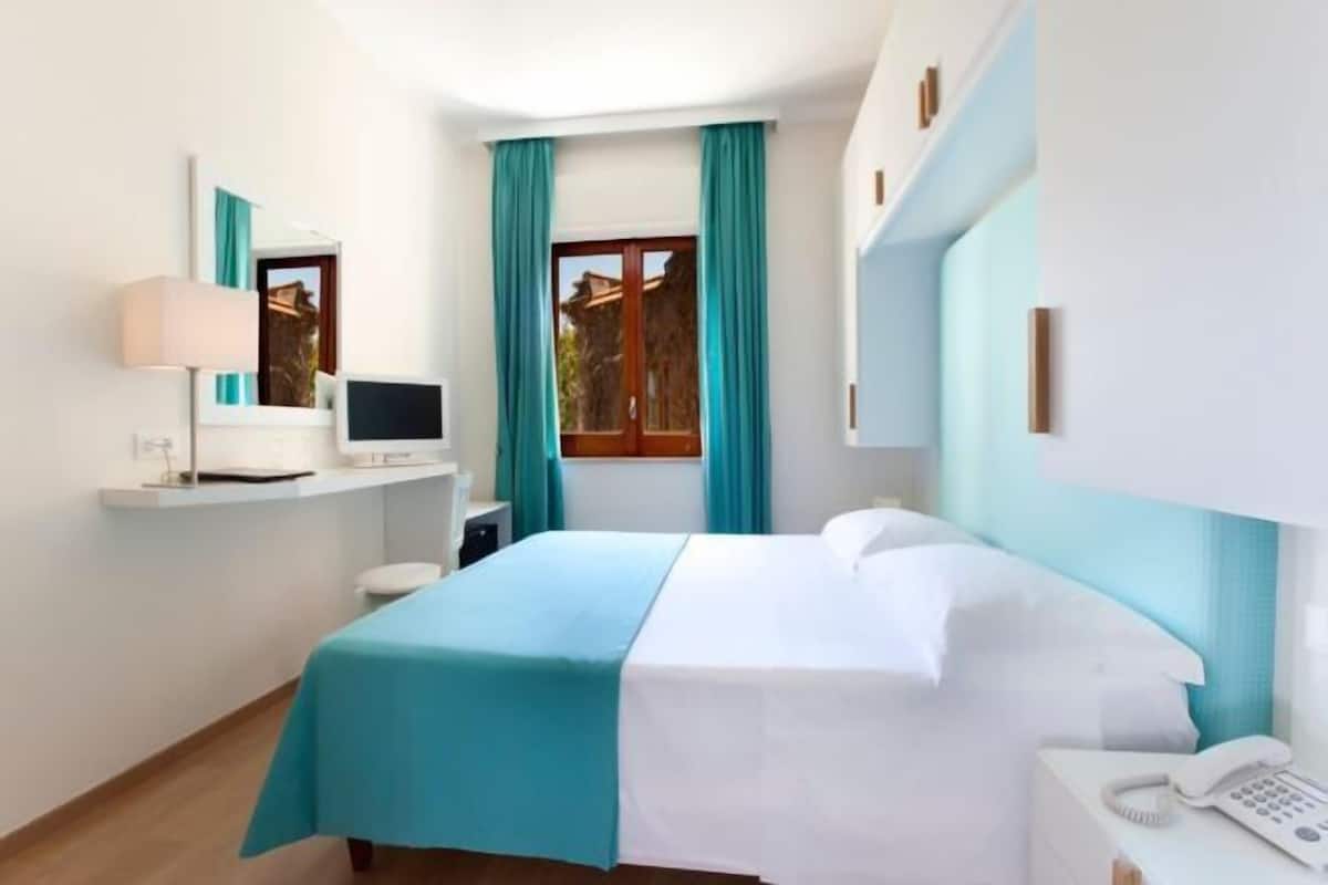 Een hotelkamer met een bed, een bureau met een computer, een telefoon en een raam met gordijnen.
