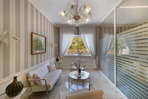 A room with a striped wall, a sofa, a table with a vase, a chandelier, and a window with curtains.