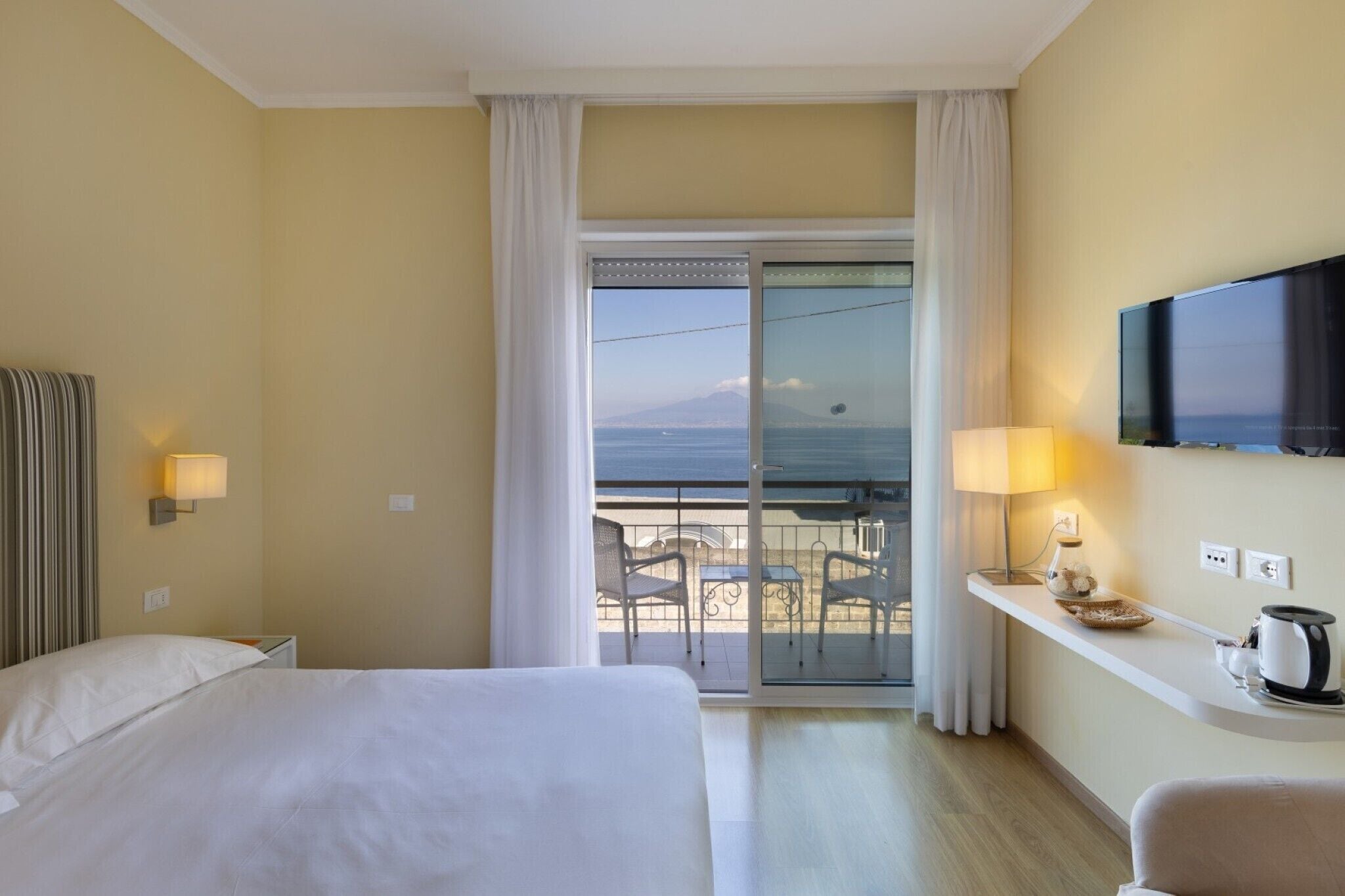 A hotel room with a bed, a TV, a balcony with a view, and a small table with a lamp.