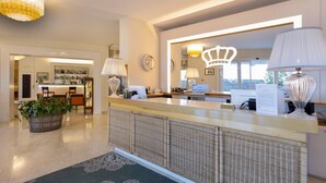 Ein Hotel-Empfangsbereich mit Theke, einer Chandelüster, einer Uhr und Blick ins Freie.