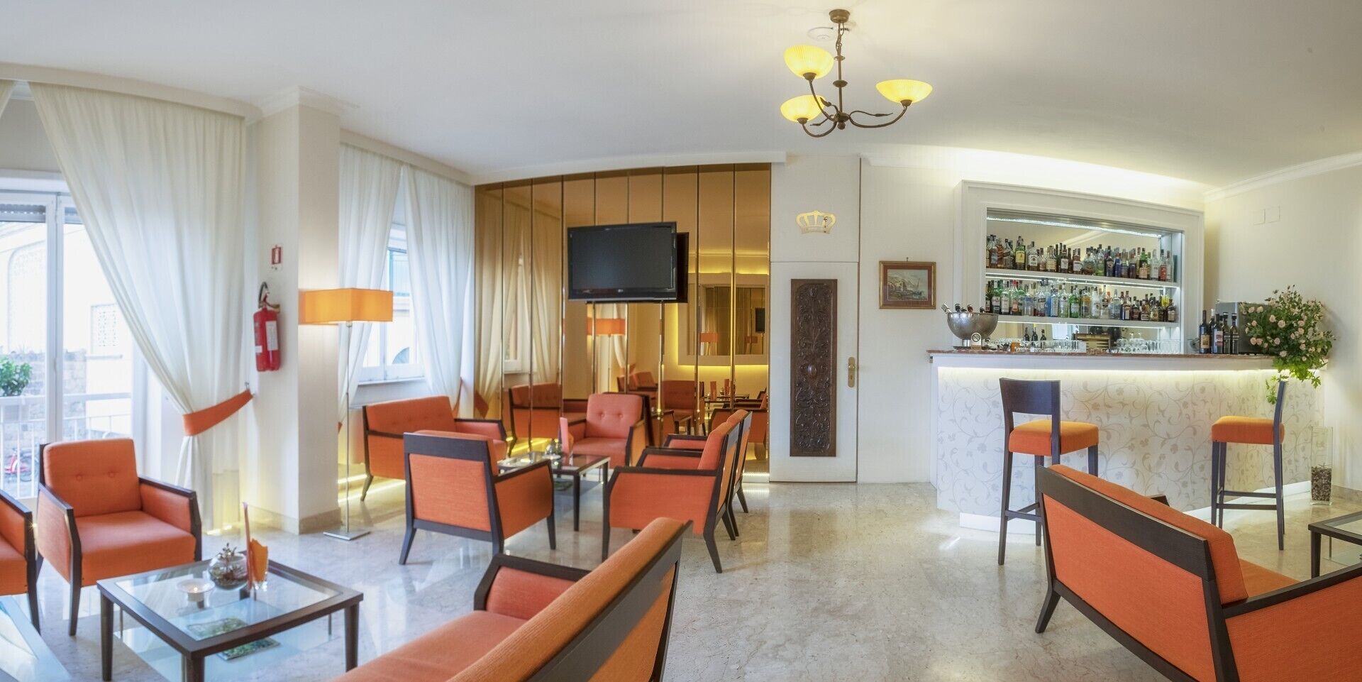 Un hall de hotel con asientos naranjas, una zona de bar, un televisor y un extintor.