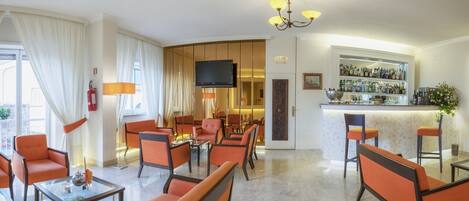 Un hall de hotel con asientos naranjas, una zona de bar, un televisor y un extintor.