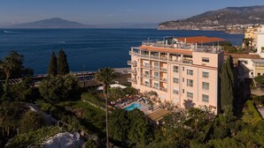Ein mehrstöckiges Hotel mit Pool, umgeben von Grün und mit Blick auf eine Küstenlandschaft sowie auf das Meer und ferne Berge.