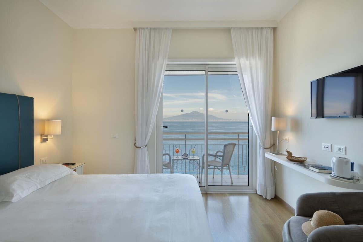 Een hotelkamer met een bed, een bureau met lamp, een televisie, een balkon met uitzicht op zee en een stoel.