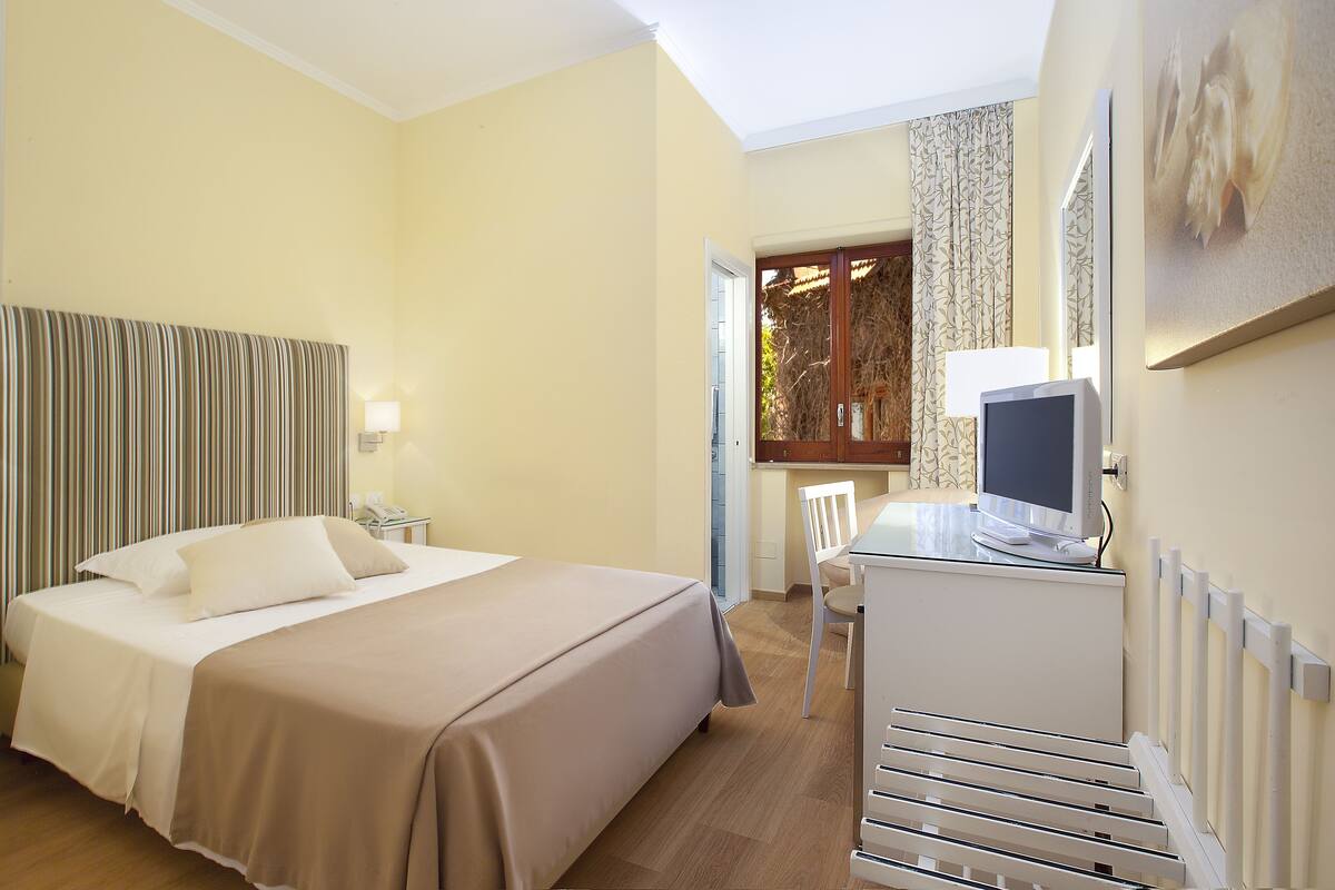Een hotelkamer met een bed, een bureau met computer, een stoel, een raam met gordijnen en een radiator.