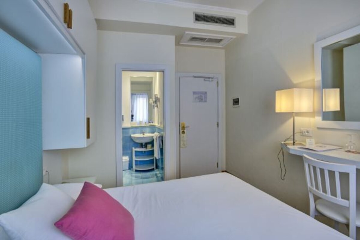 Une chambre d’hôtel comprenant un lit, une table de chevet, un miroir, une salle de bain avec un lavabo et un miroir, et une porte donnant sur une autre pièce.