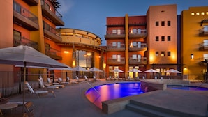 A pool area with lounge chairs and umbrellas, surrounded by multi-story buildings with balconies and outdoor lighting.