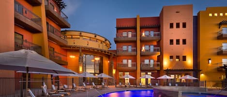 A pool area with lounge chairs and umbrellas, surrounded by multi-story buildings with balconies and outdoor lighting.