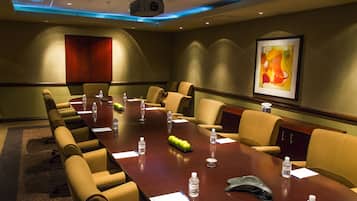 Un salón de conferencias con una larga mesa, sillas color beige, botellas de agua y un proyector.