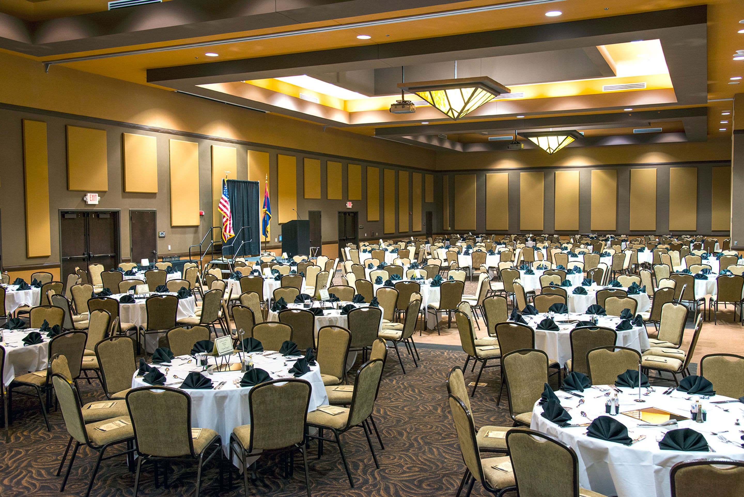 A large banquet hall with rows of tables and chairs, a stage with an American flag, and a carpeted floor.