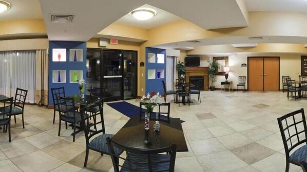 A hotel lobby with multiple dining tables and chairs, a fireplace, and a blue wall with framed pictures.