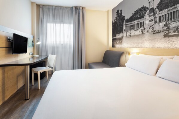 Habitación de hotel con cama, escritorio y silla, televisor y un amplio ventanal con cortinas.