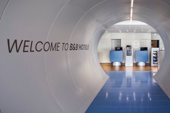 A modern hotel lobby with a curved wall displaying "WELCOME TO B&B HOTELS" and reception counters.
