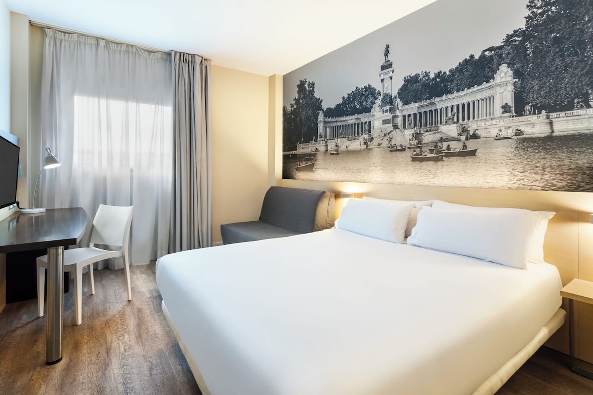 A hotel room with a large bed, a desk, a chair, and a wall-mounted TV.