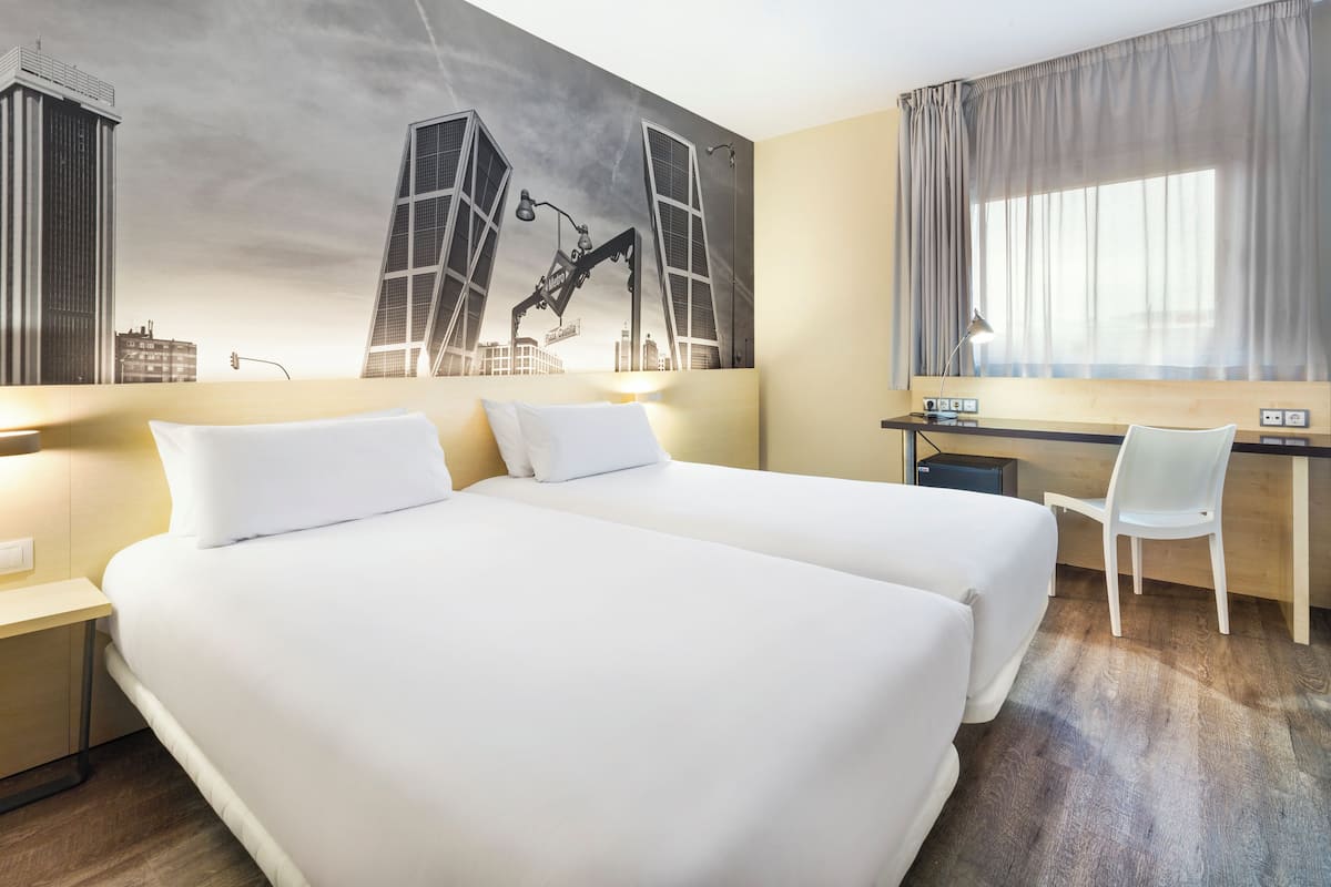 A hotel room with two beds, a desk, a chair, and a large cityscape mural on the wall.