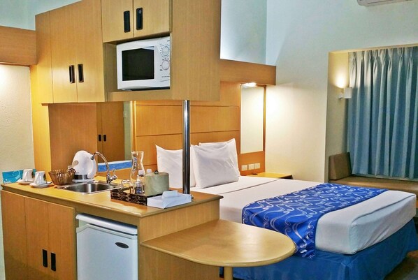 A hotel room with a kitchenette, a bed with a blue blanket, a bedside table, a lamp, and a window with curtains.