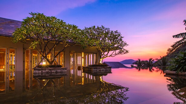 Un entorno de resort tranquilo con una piscina reflectante, un edificio bien iluminado con puertas de vidrio y vistas al atardecer.