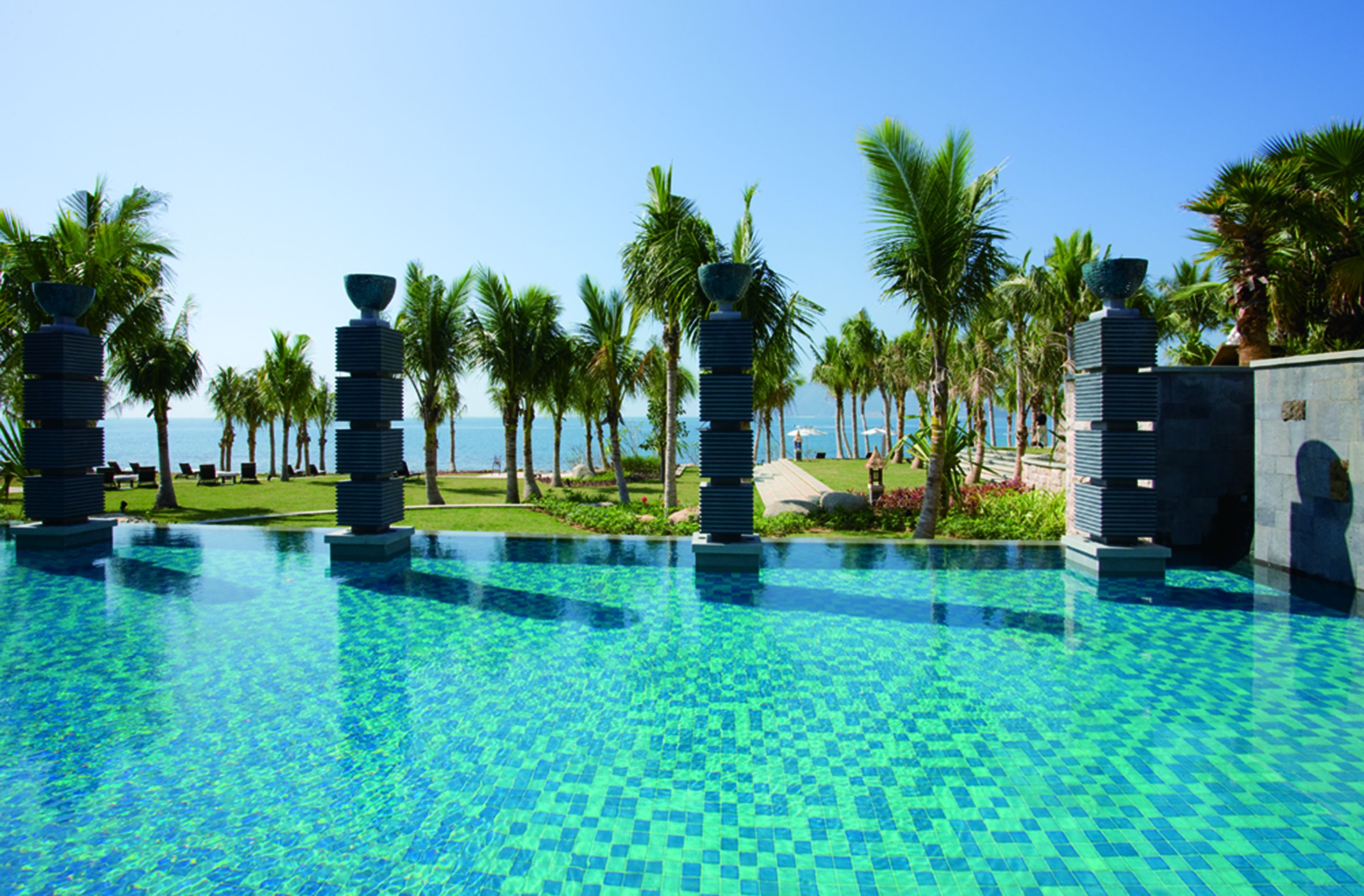 A pool with a tiled pattern, palm trees, and a view of the ocean.