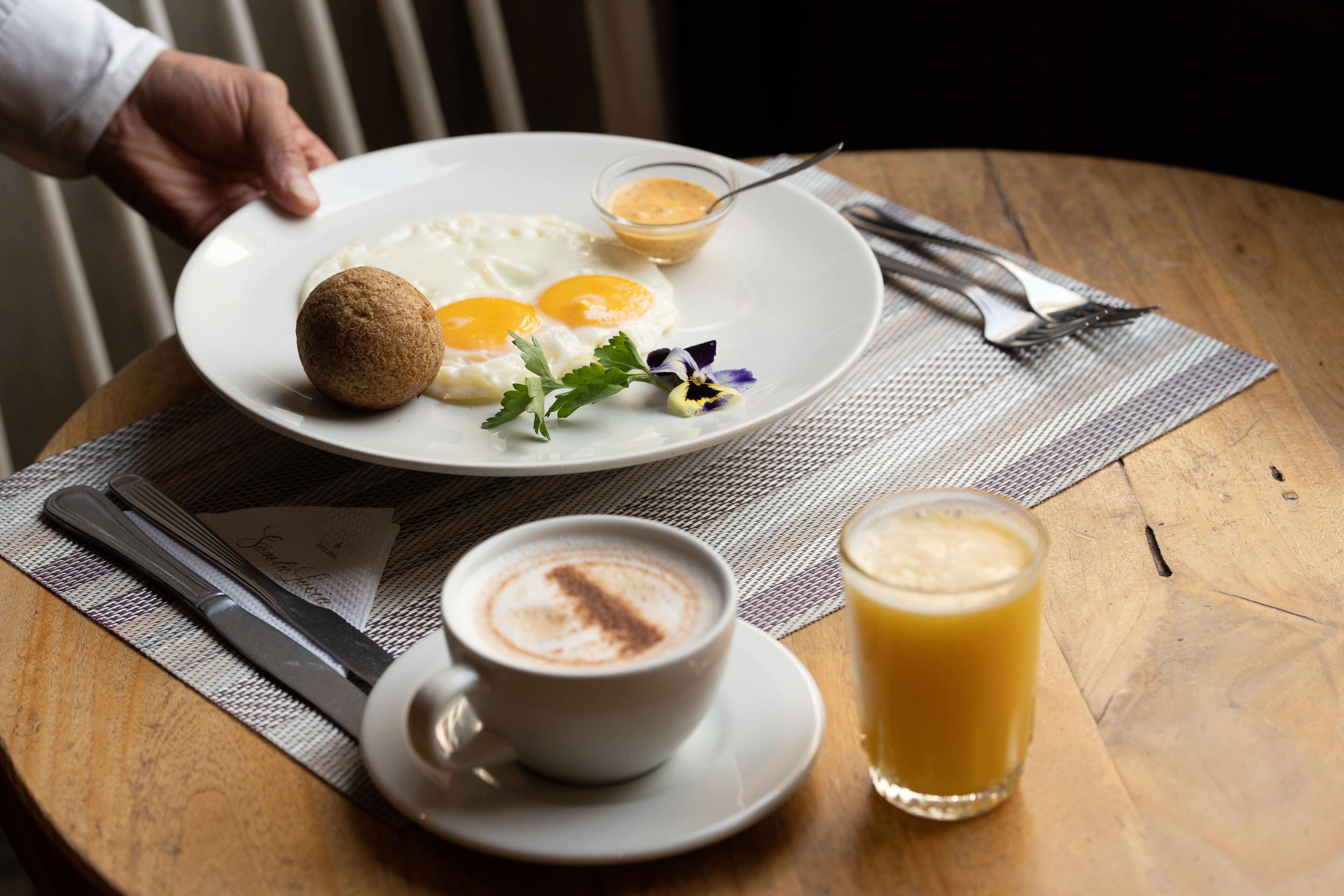 Une assiette de petit-déjeuner avec des œufs, un croissant et un petit bol de sauce, accompagnée d’une tasse de café et d’un verre de jus d’orange, le tout posé sur une table en bois.