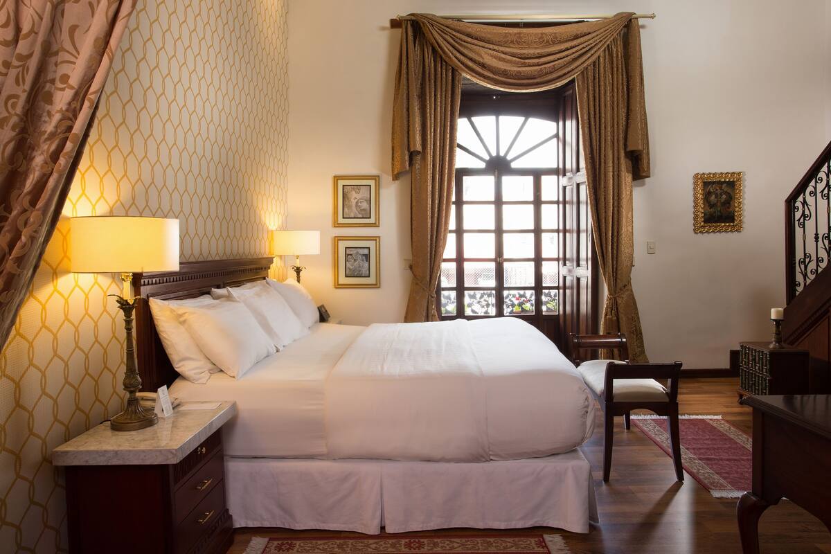 A neatly arranged bedroom with a large bed, bedside lamps, a chair, and a desk with books.