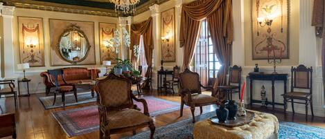 A grand room with ornate chandeliers, large windows with draped curtains, and classic furniture.