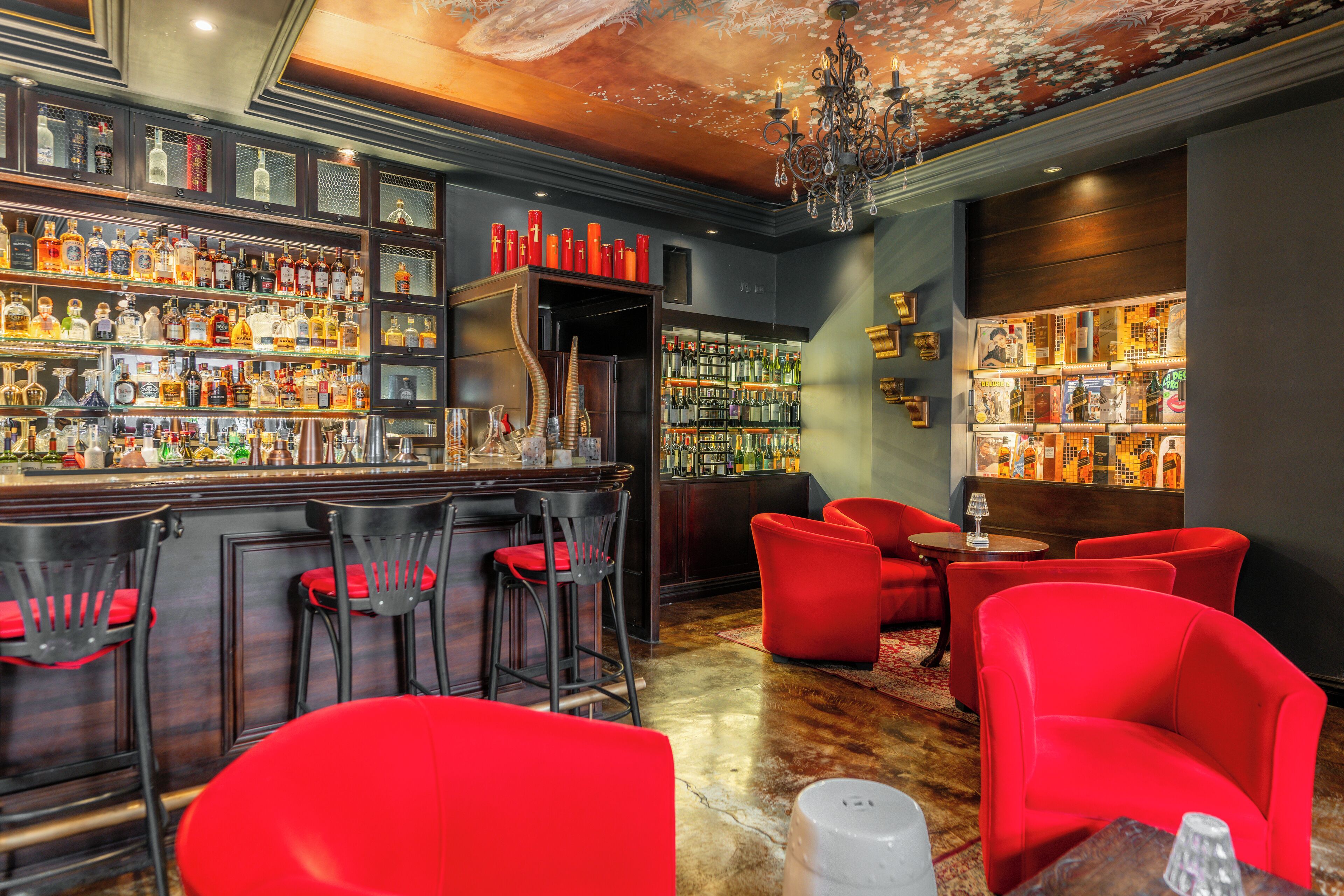 A bar area with multiple shelves of liquor bottles, red upholstered chairs, and a bar counter with stools.