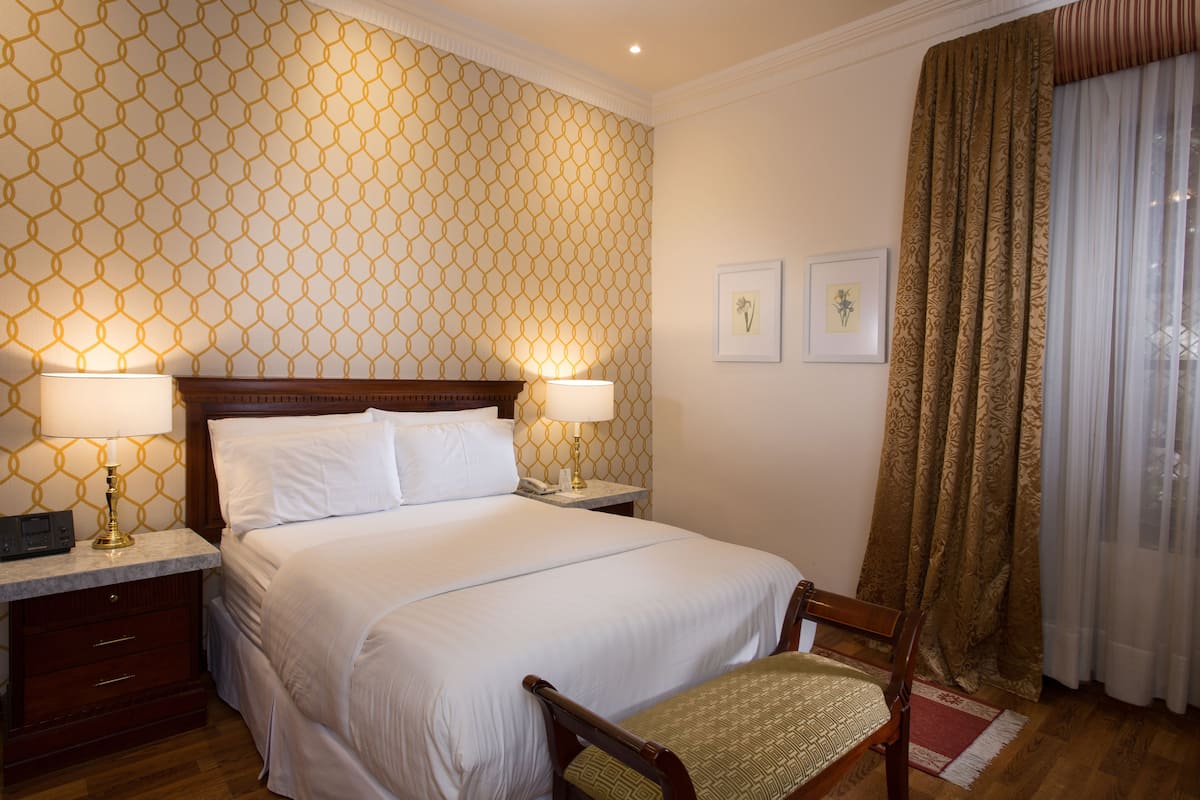 A neatly made bed with white linens, two bedside lamps, a wooden nightstand, and a chair with a patterned cushion.