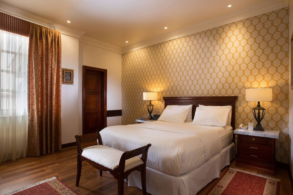 A neatly arranged hotel room with a large bed, two bedside tables with lamps, a wooden chair, and a patterned wall.