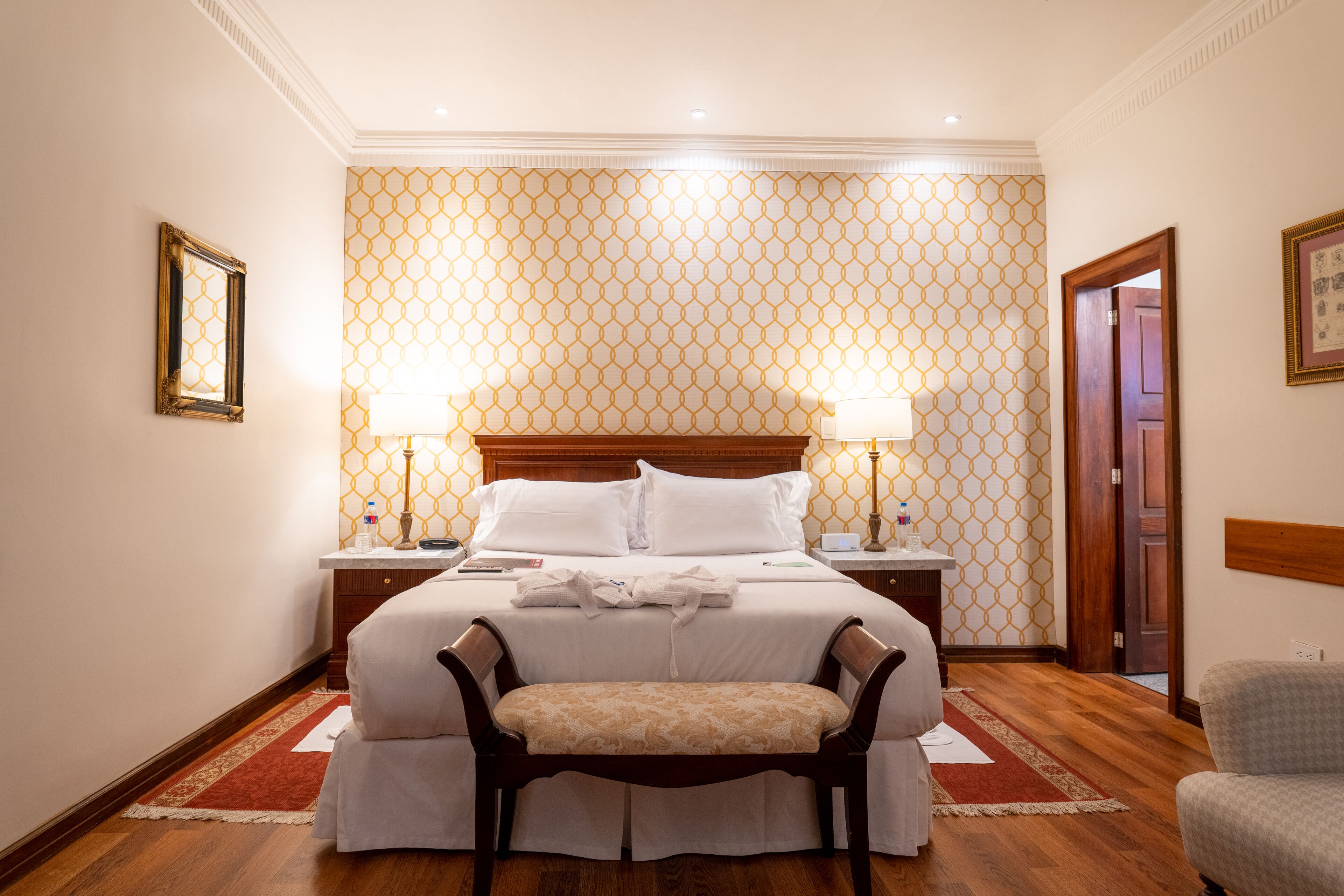 A hotel room with a large bed, two bedside tables, a wooden bench, and a patterned wallpaper.
