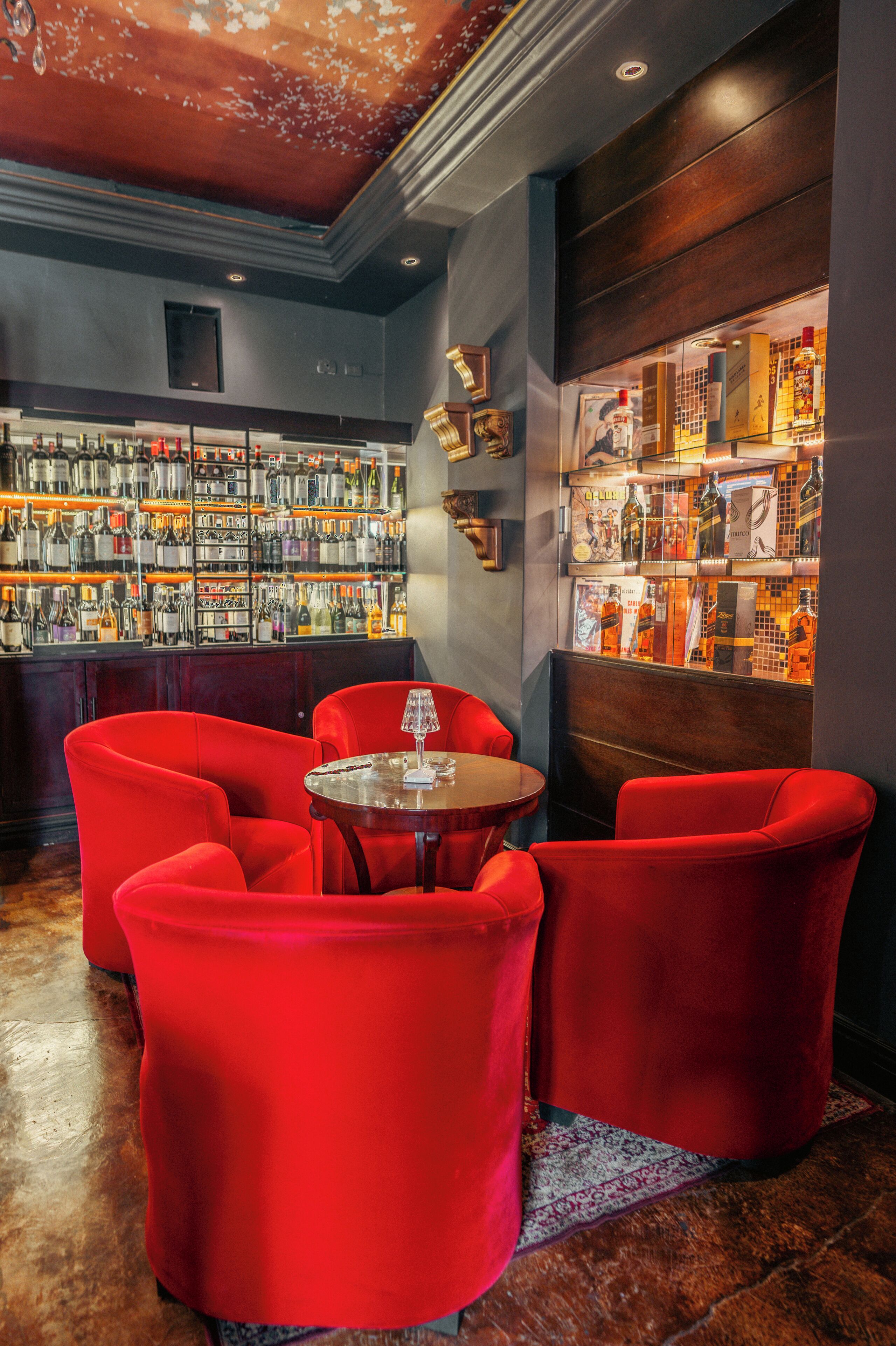 A bar area with red upholstered chairs, a round table with a wine glass, and shelves stocked with liquor bottles.