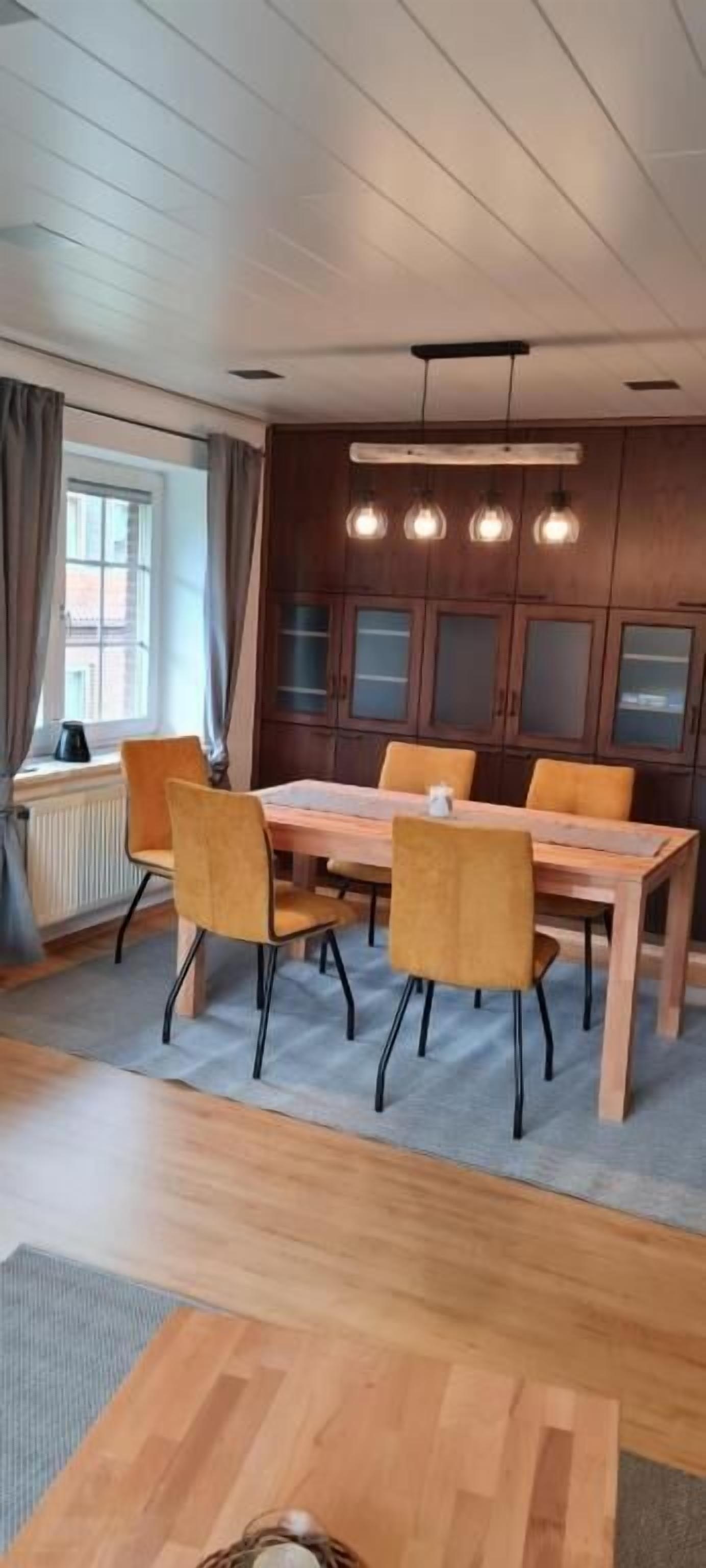 Ruokailutila, jossa on puinen pöytä ja tuolit, ikkuna, jossa on verhot, sekä puiset kaapit lasiovilla.